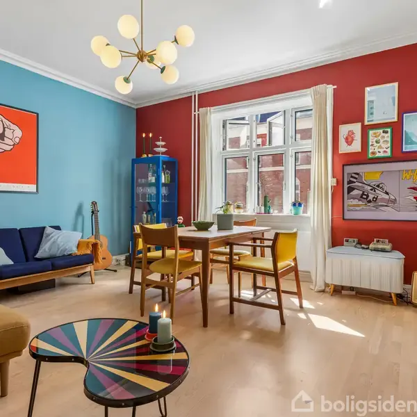 Stue med farverige vægge, blå og rød, møbleret med sofa, borde og stole. Vægkunst inkluderer en knyttet næve og et billede af en bil. Midt i rummet hænger en moderne lysekrone.