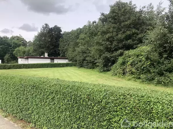 Huset står stille mellem en velplejet hæk og frodig skov, omgivet af en stor græsplæne under en overskyet himmel.