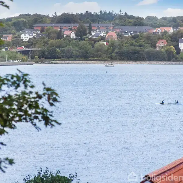To personer padler i kajakker på en rolig sø omgivet af grønne træer og en landsby med farverige bygninger i baggrunden.