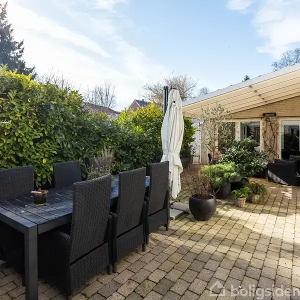 Langt havebord med stole står i solrig terrasse omgivet af grønne planter og buske. I baggrunden ses en murstensbygning med store vinduer og en overdækket veranda.