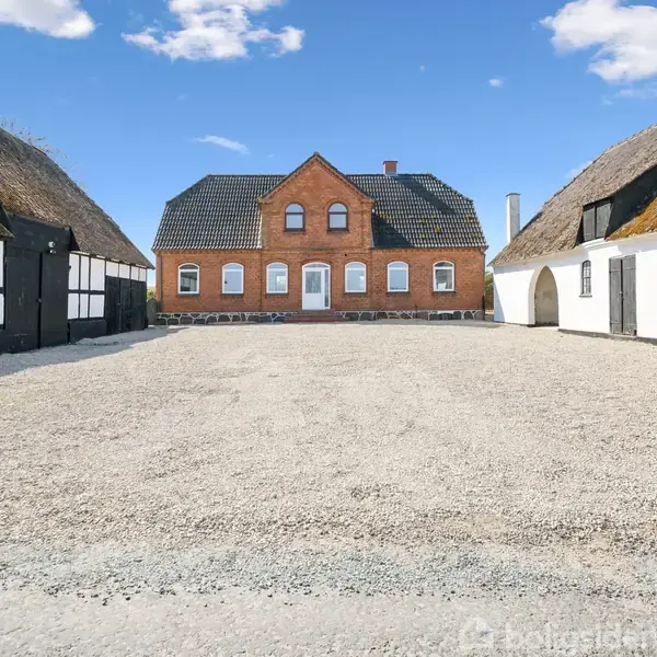 En rød murstensbygning står centralt i en gårdsplads, omgivet af bindingsværkshuse med stråtag under en blå himmel med skyer.