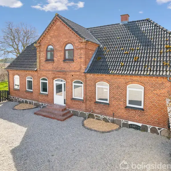 Rødt murstenshus står stillet mod en grusbelagt gårdsplads, omgivet af yderligere bygninger. Huset har karakteristiske buede vinduer og et sort tegltag. Baggrund viser åbne marker og en stor, bar gren.