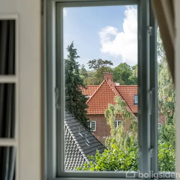 Et åbent vindue indrammer et rødt tegltag og træer udenfor; himlen er blå med lette skyer.
