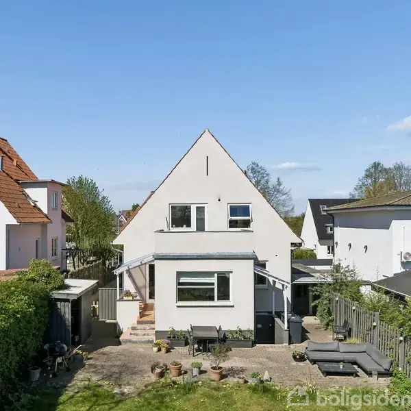 En hvid to-etagers villa med skråt tag står stille i en velplejet have. Omkring huset er der andre villaer og grønne buske. Jardiner og en terrasse ses foran huset.