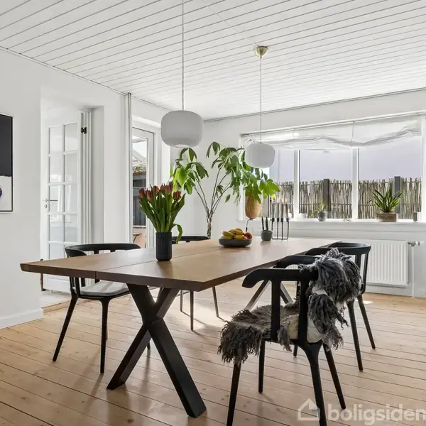 Spisebord med stole står i et lyst rum. Vaser med blomster og frugtskål pynter bordet. Store vinduer bagved slipper meget lys ind, og en moderne lampe hænger over bordet.