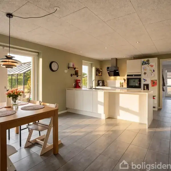 Et lyst køkken med åbne hylder og moderne apparater. Et spisebord med stole står ved et vindue, der åbner til en terrasse. Blomster i en vase pynter bordet.
