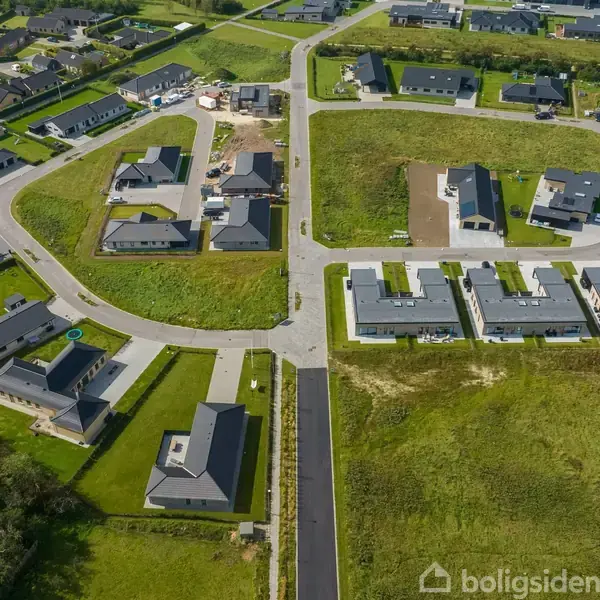 Boligområde med moderne huse arrangeret omkring åbne grønne områder. En vej løber gennem landskabet, og husene er placeret i grupper på begge sider, omringet af græs og natur.
