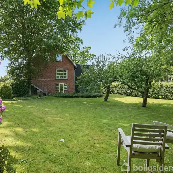 Et par hvide træstole står stille på en grøn græsplæne. I baggrunden ses et rødt murstenshus omgivet af høje træer og buske i en solrig have.