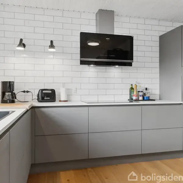 Gråt køkken med moderne apparater, herunder kaffemaskine og ovn. Hvide fliser på væggen og trælaminatgulv. To væglamper lyser rummet op. Bordpladen er hvid og minimalistisk indrettet.
