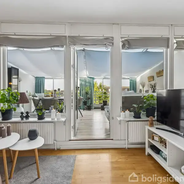 En stue med sofabord er centreret omkring en stor skærm. Glasdøre åbner til en lys, grøn beplantet udestue. Hylder og radiator er dekoreret med planter.