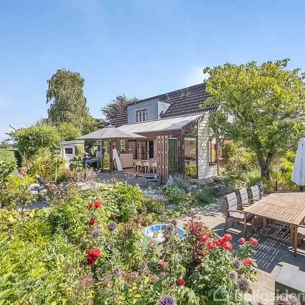 Hus med terrasse, omgivet af frodige planter og blomster. Terrasse med stort træbord og paraply. Bagved ses marker og en blå himmel.
