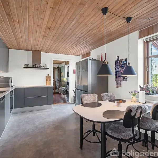 Et moderne køkken med grå kabinetter og træloft. Et rundt spisebord med fire stole står under to loftlamper. Store vinduer giver udsigt til en grøn have.
