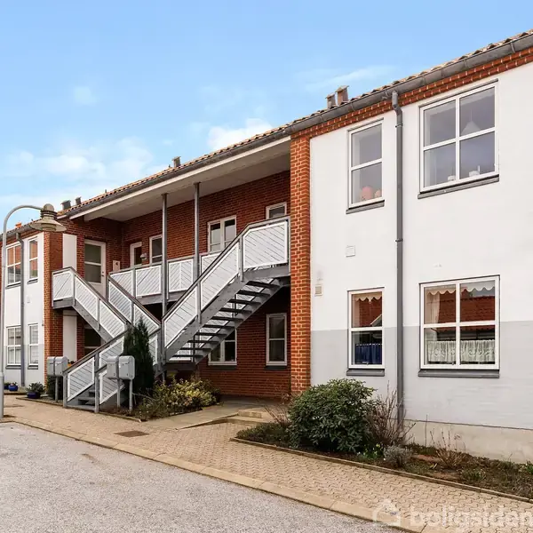 En række af toetages murstensrækkehuse med udendørs trapper forbinder første og anden etage. En bil holder parkeret i en asfalteret gårdsplads med planter langs facaden.