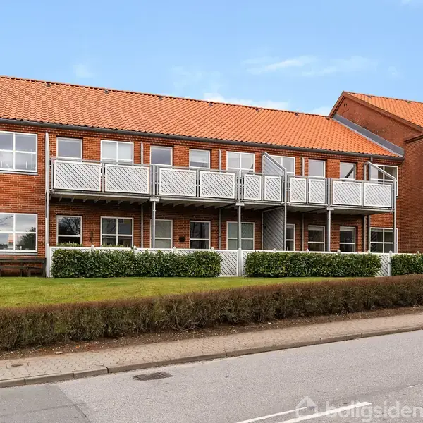 Rækkehus med rødt tegltag og altaner strækker sig langs en rolig vej. Grønt buskads omgiver ejendommen, der står under en klar blå himmel.