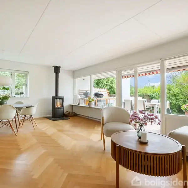 Stue med en brændeovn, lyse trægulve og store vinduer. Udenfor ses en terrasse med udsigt over en frodig have. Møblerne er moderne og minimalistiske.