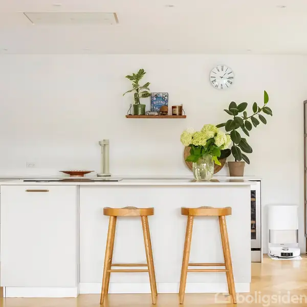 To træstole står ved en moderne køkkenø med hvide skabe. En vase med blomster står ovenpå. Bagved er en hylde med planter og et ur på en hvid væg.