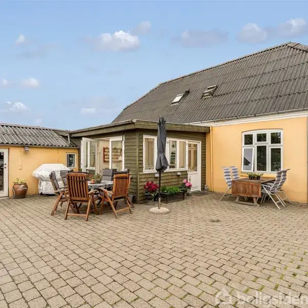 Huset har en gul facade med gråt tag. På terrassen står havemøbler og en parasol. Murstensbelægning omgiver området, mens krukker med planter tilføjer dekoration.