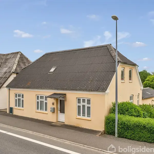 Et gulmalet hus med mørkt tag ligger langs en vej med fortov. Huset har hvide vinduesrammer, en lille indgangsforplads og buske i haven. Himlen er blå med få skyer.