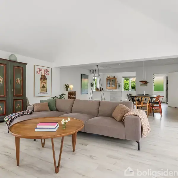 En stue indeholder en brun sofa og et rundt sofabord med bøger og blomster. I baggrunden ses et spiseområde. På væggen hænger en plakat med teksten "TUDSEN PÅ FØJEL".