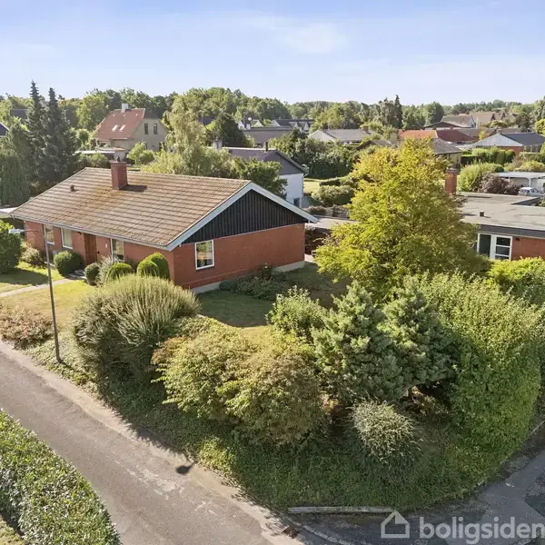 Et rødt murstenshus med skråtag omkranset af velplejede buske og træer. En bil er parkeret ved siden af huset. Det er beliggende i et kvarter med flere huse omkring.