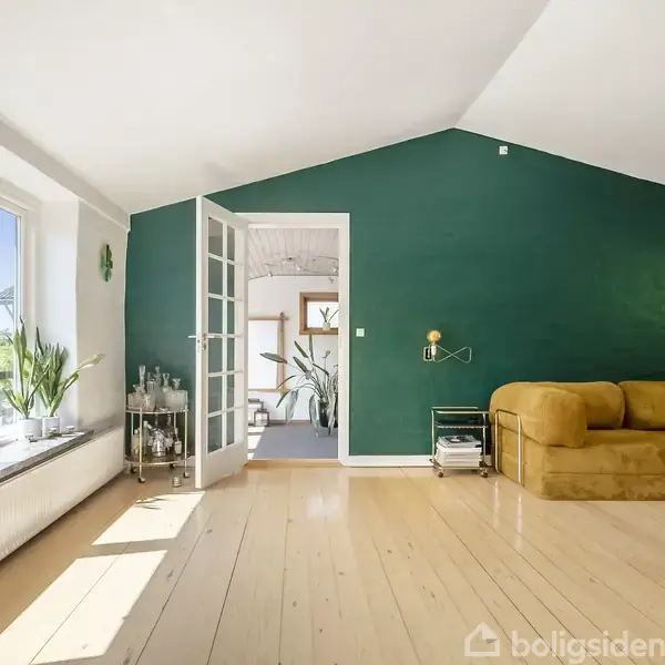 Brun sofa står i hjørnet af et rum med en grøn væg. Store vinduer til venstre viser et solrigt landskab. Åbne franske døre fører til et tilstødende rum.