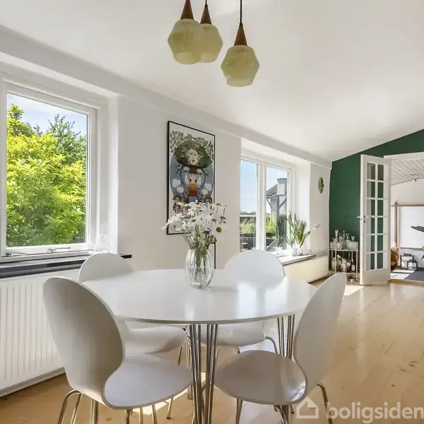 Rundt bord med hvide stole står under tre hængende lamper; en vase med blomster pryder bordet. Store vinduer slipper dagslys ind i det lyse rum med grøn væg i baggrunden.