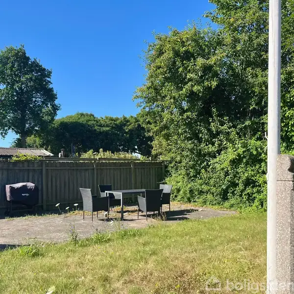 Terrassemøbler står stille på en flisebelagt patio med fire stole omkring et bord. Omkring patioen er en græsplæne og tætbevokset hæk, og i baggrunden ses en træbeklædt hegn og en stor træ.