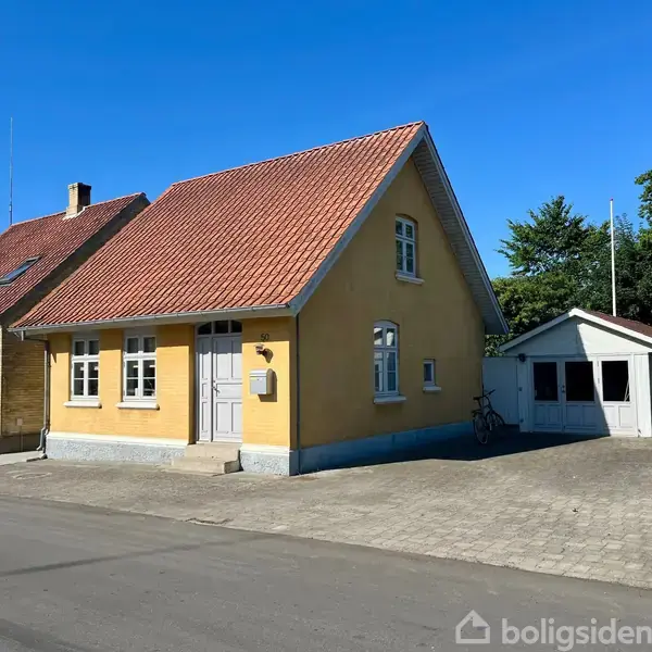 Et gulmalet hus med rødt tegltag står stille ved en stille vej. Det er omgivet af en garage og indkørsel, mens træer i baggrunden antyder et roligt kvarter.