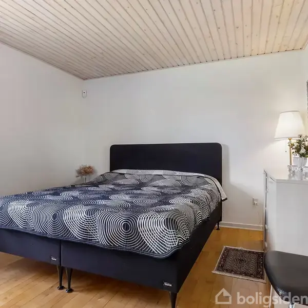 En dobbeltseng med geometrisk mønstret sengetæppe står i et soveværelse. En kommode med lampe og blomster er ved siden af, omgivet af lyse trævægge og gulve.