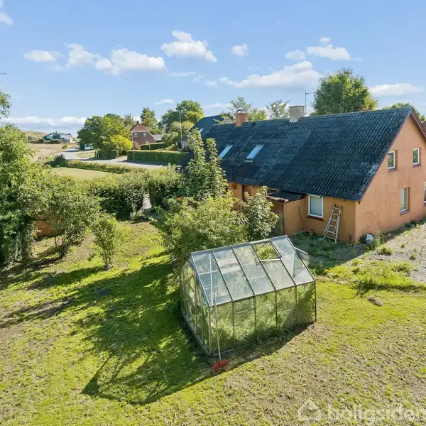 Et grønt drivhus står i en solrig have. En orange murstensbygning med sort tag ses bagved, omgivet af træer og buske. Skyer spotter den klare blå himmel.