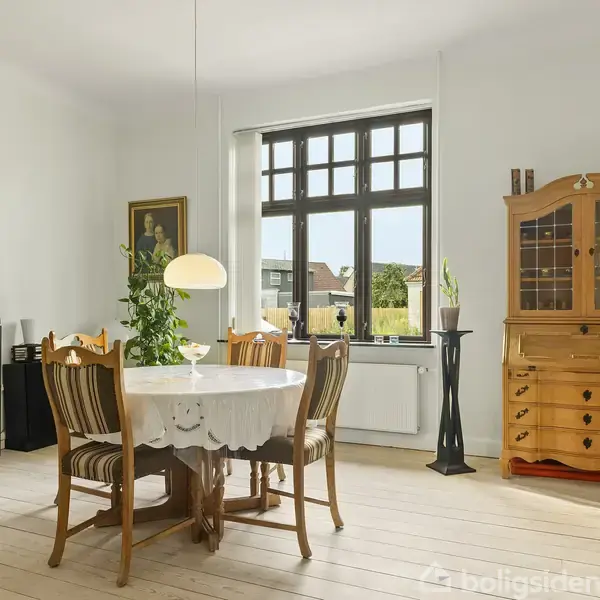 Et spisebord med dug og stole står i centrum af et lyst rum. Bagved ses en træbuffet og et stort vindue med udsigt til en have. Ingen tekst fundet.