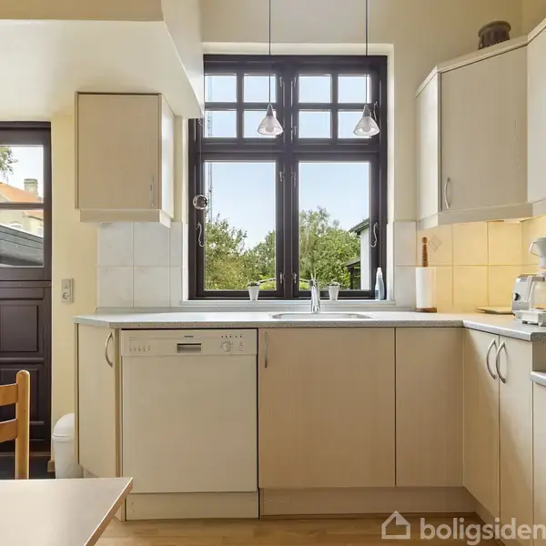 Et lyst køkken med hvidt interiør; opvaskemaskine og kaffemaskine står på en køkkenbord. Store vinduer giver udsigt til grønt udenfor. En dør fører til en have.