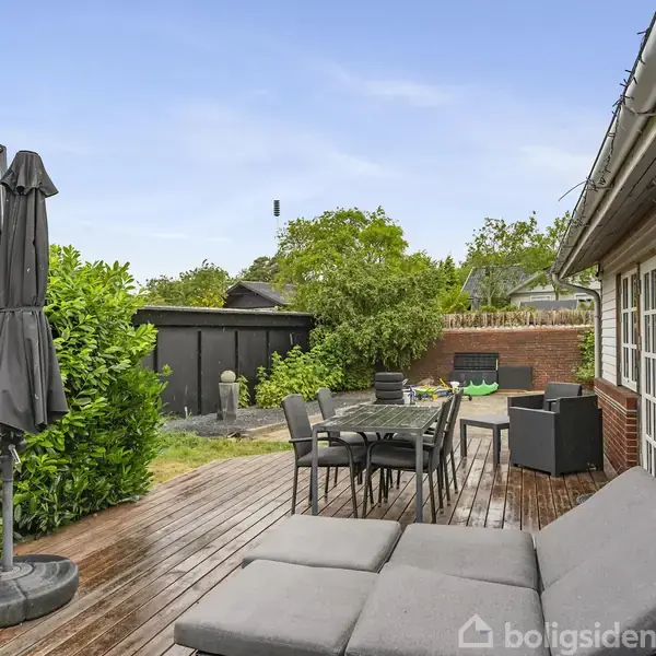 Terrasse med grå solsenge og sort parasol. Et udendørs borde-sæt er placeret ved siden af et hus, omgivet af buske og et skur. Atmosfæren er afslappende og velplejet.