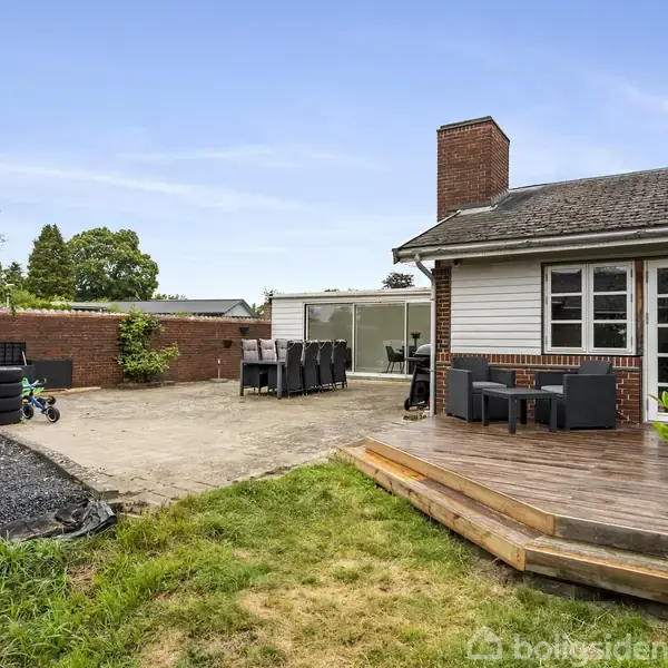 Hus med terrasse og skydedør fører ud til gårdhave omgivet af grønne træer og græsplæne. Der er udendørs møbler og en baggård med murstensvæg og legetøj.