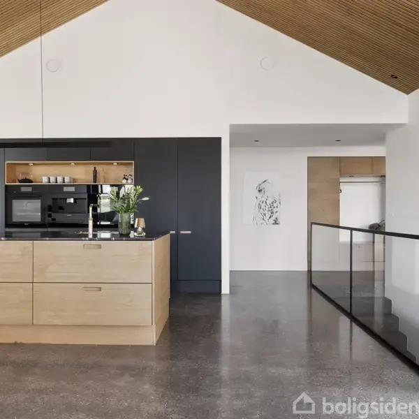 En moderne køkkenø i lyst træ står i forgrunden. Bagved ses sorte skabe med indbyggede apparater. Loftet er højt og af træ, og gulvet er poleret beton.