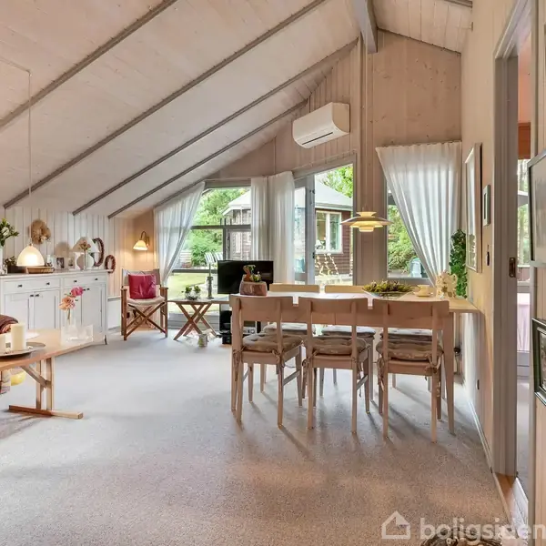 Stue med skrå loft, hvor spisebord med seks stole står ved vinduer. Sofa og bord er til venstre. Væggene har billeder og vinduer med lyse gardiner. Ingen tekst findes.