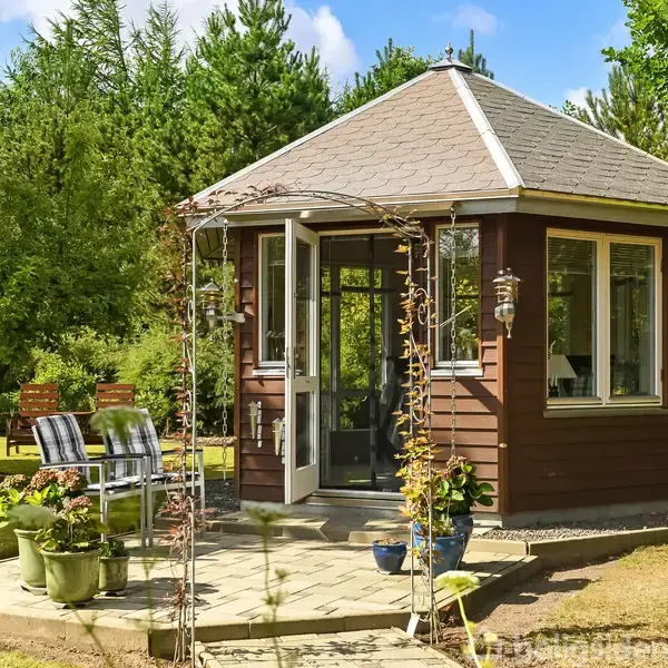 En lille, brun havepavillon står midt i en frodig have med græsplæne og træer. Foran pavillonen er der terrassestole og blomsterkrukker arrangeret på en flisebelagt sti.