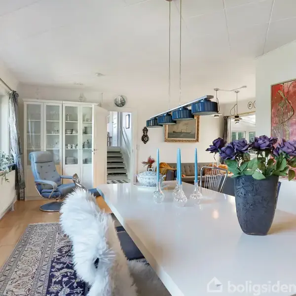 Et langt spisebord er dækket med stel og blå lys i en lys spisestue. En vase med lilla blomster står midt på bordet. Rummet har kunst på væggene og et vitrineskab.