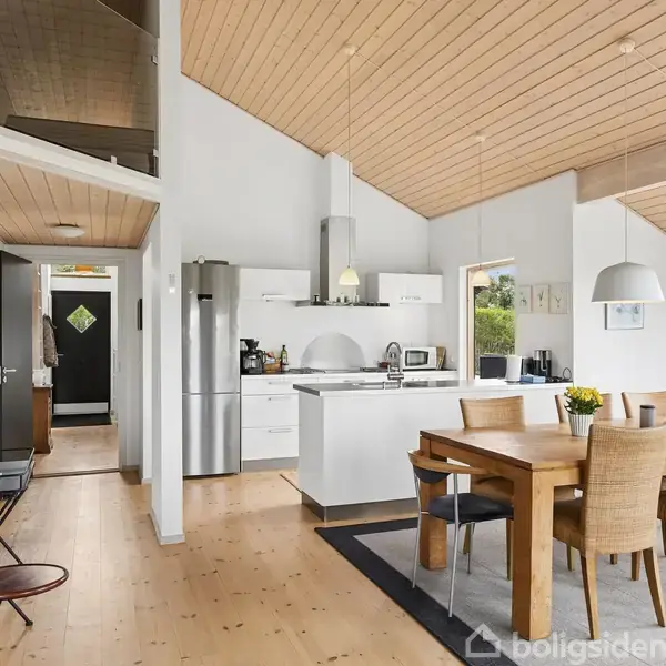 Køkken-alrum med spisebord i træ og seks stole. I baggrunden ses et moderne køkken med hvidt interiør og stål køleskab. Store vinduer giver udsigt til grønt område udenfor.