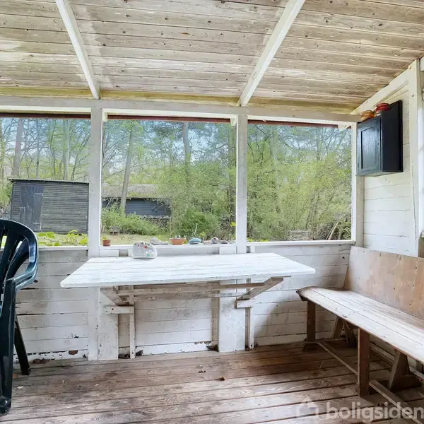 Et træbord står på en veranda med en plastikstol og en lang træbænk. Omgivet af hvide vægge og træloft, ses en skov gennem vinduerne.