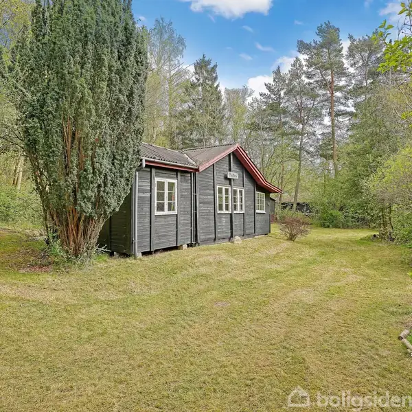 Et mørkt træhus står stille omgivet af en frodig have og høje træer. Græsplænen strækker sig bredt, mens himlen over er klar med få skyer.