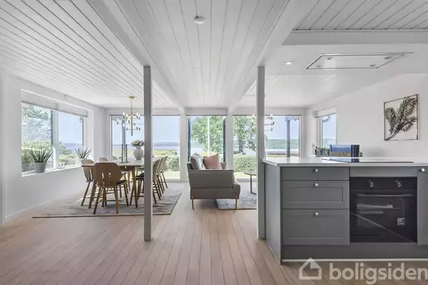 Stue med spisebord og stole ved store vinduer, der giver udsigt til have og sø. Stort gråt køkkenbord i forgrunden under hvidt træloft.