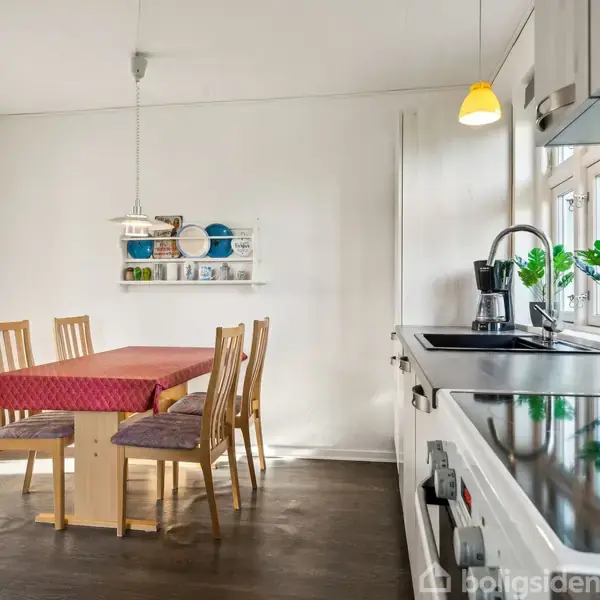 En spisebord med fire træstole, dækket med en rød dug, står i et lyst køkken. En hylde med tallerkener hænger på væggen, og et køkkenbord med planter er til højre.