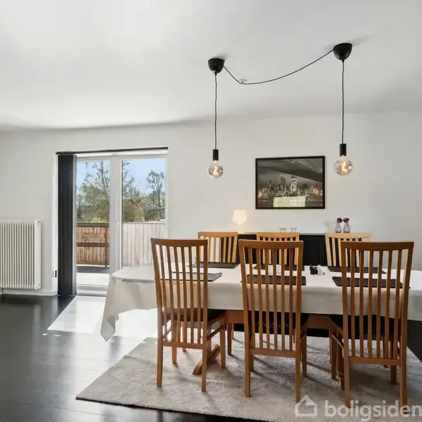 Spisebord med seks træstole dækket med hvid dug står i et lyst spisestueområde. To hængelamper over bordet. Væggen har kunstværker, og en dør fører ud til en terrasse.