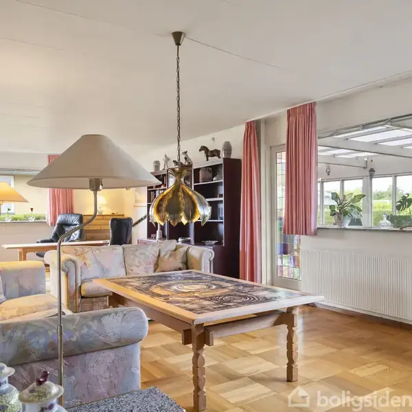 En stue med et blomstret sofaarrangement omkring et træbord. En hængelampe lyser op i rummet. Store vinduer med lyserøde gardiner åbner op til en lys udestue med planter.