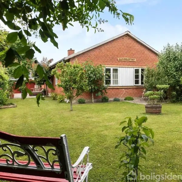 Rød murstenshus med teksten "SANDMOLLEN", omgivet af en grøn have med træer og buske; en rød-hvid bænk står i forgrunden.
