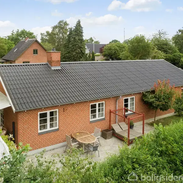 En rød murstensbygning med sort tag står omgivet af frodige buske og træer. Foran bygningen er en stenlagt terrasse med et bord og stolearrangement.