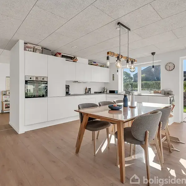 Spisebord med stole står i et lyst køkken/alrum med trægulv. Moderne lysarmatur hænger over bordet. Køkkenet har indbyggede ovne og store vinduer, der slipper naturligt lys ind fra haven.