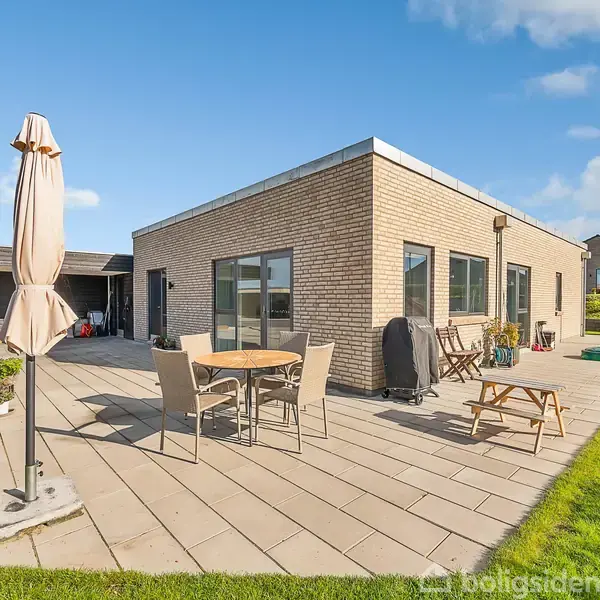 Firkantet murstenshus med store vinduer står på en flisebelagt terrasse. Haven er velplejet med græs, parasol og havemøbler. Et skur ses i baggrunden under en lys, blå himmel.
