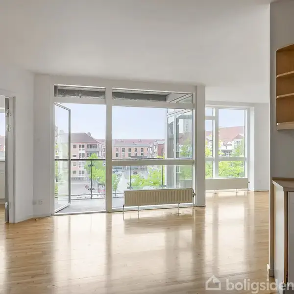 Et lyst rum med trægulv indeholder store glasdøre, som åbnes mod en balkon. I baggrunden ses bybygninger med røde tage. Højre side har lyse træhylder.